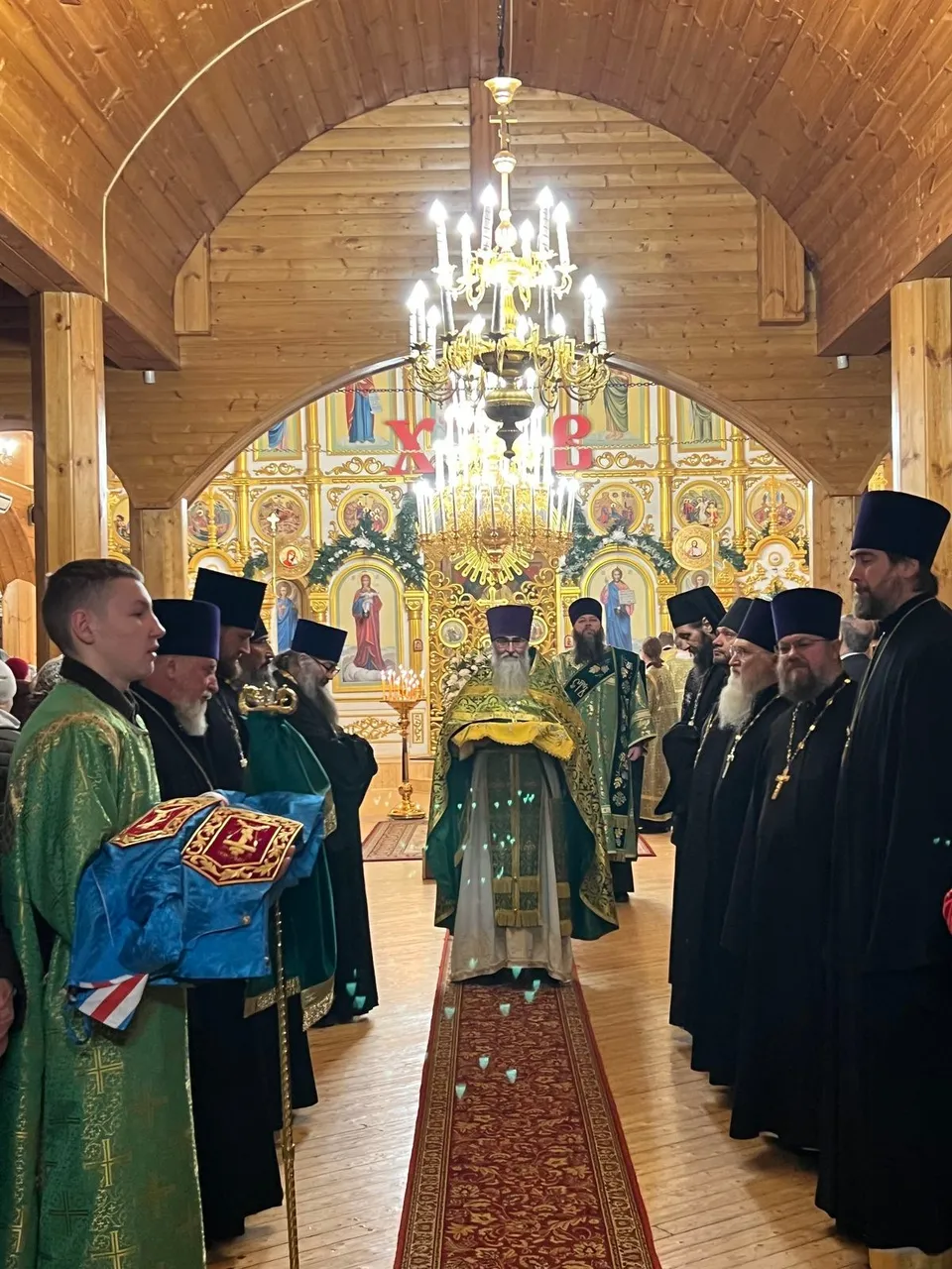 Митрополит Филарет идёт по красной ковровой дорожке к иконостасу, украшенному к празднику; рядом иподиакон с ковчежцем и духовенство