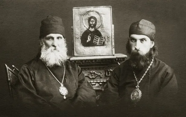 Чёрно-белый фотопортрет двух пожилых священнослужителей в монашеских скуфьях с наперсными крестами, между ними на подставке — икона Спасителя