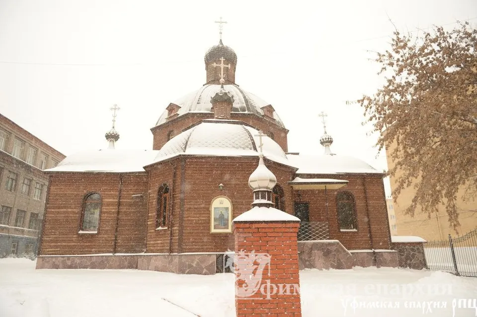 Деревянная церковь с одним большим тёмным куполом и несколькими маленькими главками, охристые бревенчатые стены, идёт снег, рядом кирпичный столб ограды с киотом; людей на фото нет