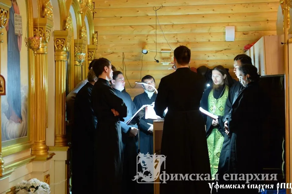 Группа певчих в чёрных подрясниках стоит у нотных папок вокруг регента, спиной к камере регент дирижирует; рядом диакон в зелёном облачении
