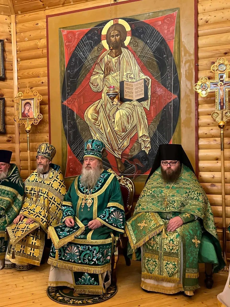 Правящий архиерей в зелёном облачении сидит между двумя священнослужителями на фоне большого живописного образа Христа на троне