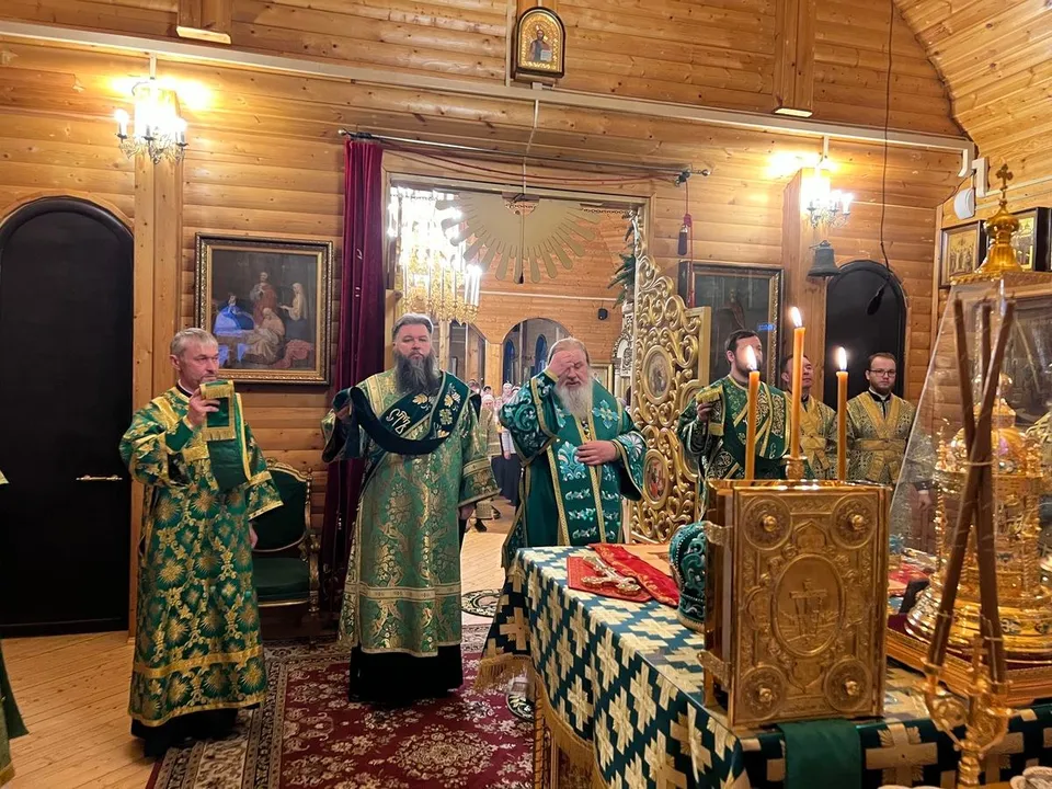 Несколько священнослужителей в зелёных облачениях стоят у престола с горящими свечами, на заднем плане — арка и паникадило